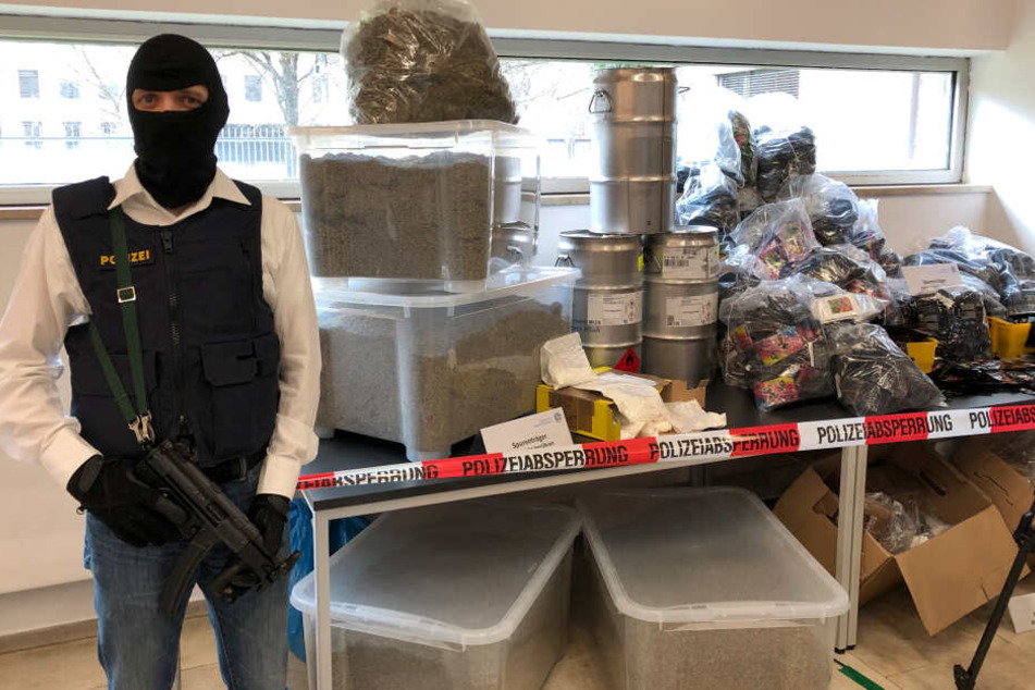 Ein bewaffneter Polizeibeamter steht im bayerischen Kriminalamt neben der Präsentation einer Drogenproduktionsstrecke und einiger Luxusprodukte aus der Wohnung eines Drogenproduzenten in München.