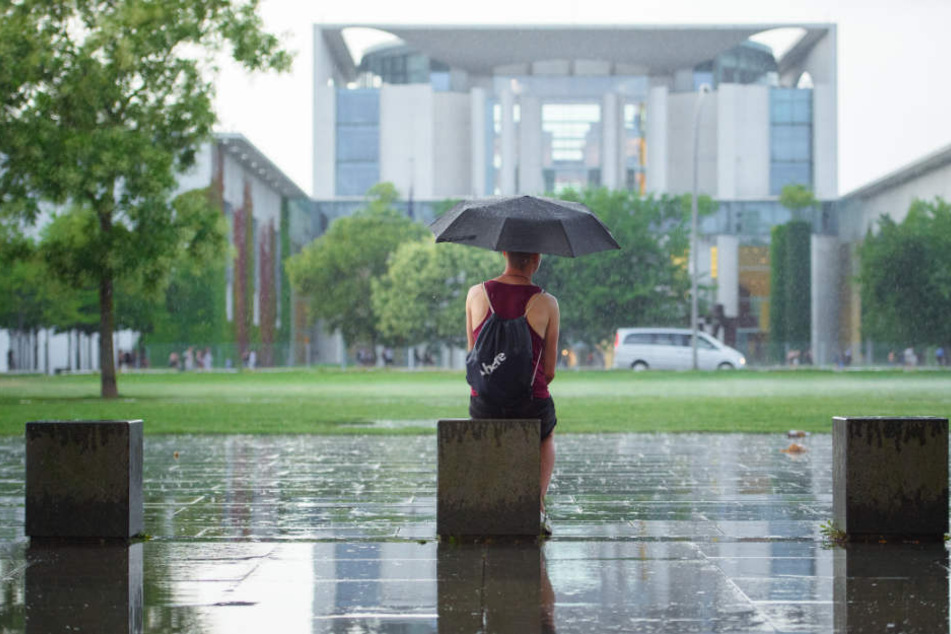 Eine Frau sitzt mit einem Regenschirm im Regen vor dem Bundeskanzleramt.