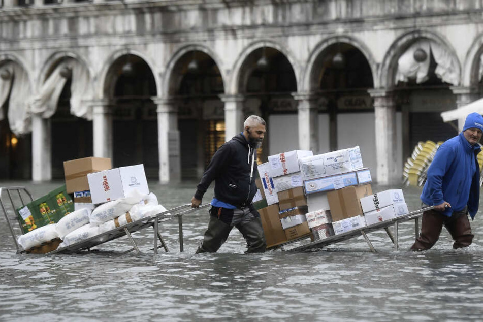 Männer waten mit Kartons und Lebensmitteln, die sie auf einer Trage transportieren, durch das Hochwasser auf einem überfluteten Platz.