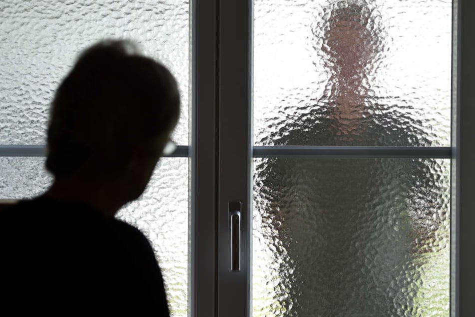 Eine Frau steht an einer Glasscheibe, hinter der ein Stalker steht. (Symbolbild)