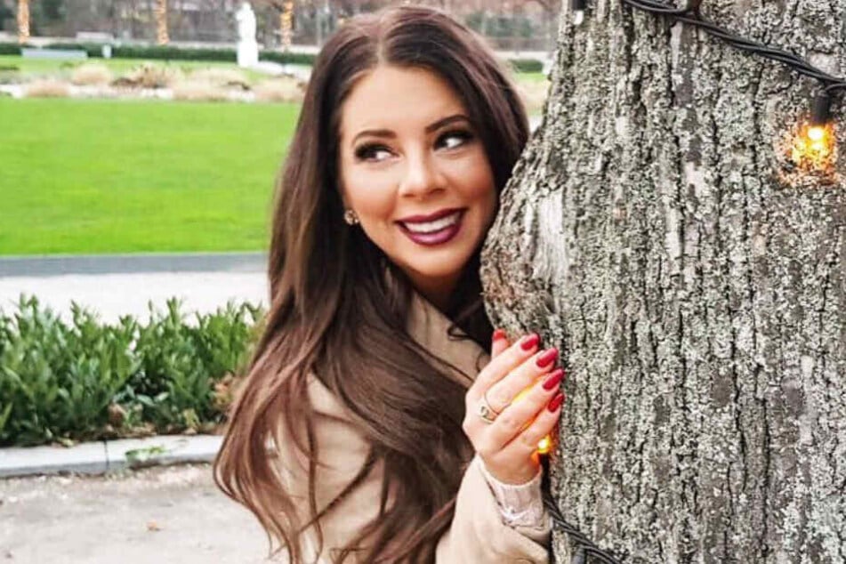 Jenny Frankhauser versteckt sich lächelnd hinter einem Baum.