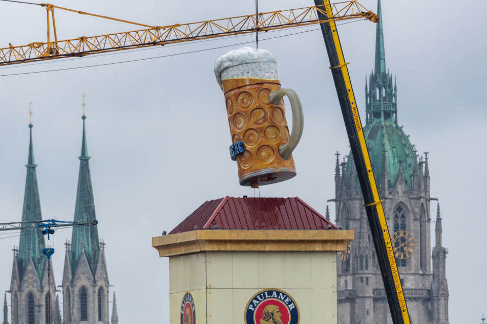 Ein übergroßer und rund rund 1,5 Tonnen schwerer Bierkrug wird mit einem Kran auf der Theresienwiese an seinen Platz gehoben. Im Hintergrund ist die Sankt Paul Kirche zu sehen.