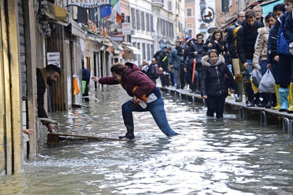 Hochwasseralarm in Venedig! Eine Frau versucht, eine überflutete Straße zu überqueren, während Menschen auf einer Gerüstbrücke laufen.