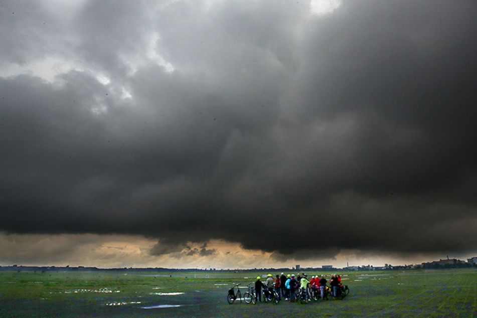 Eine Gruppe Radfahrer steht auf dem Gelände des ehemaligen Flughafens Tempelhof in Berlin, während sich über ihnen dunkle Regenwolken zusammen brauen.