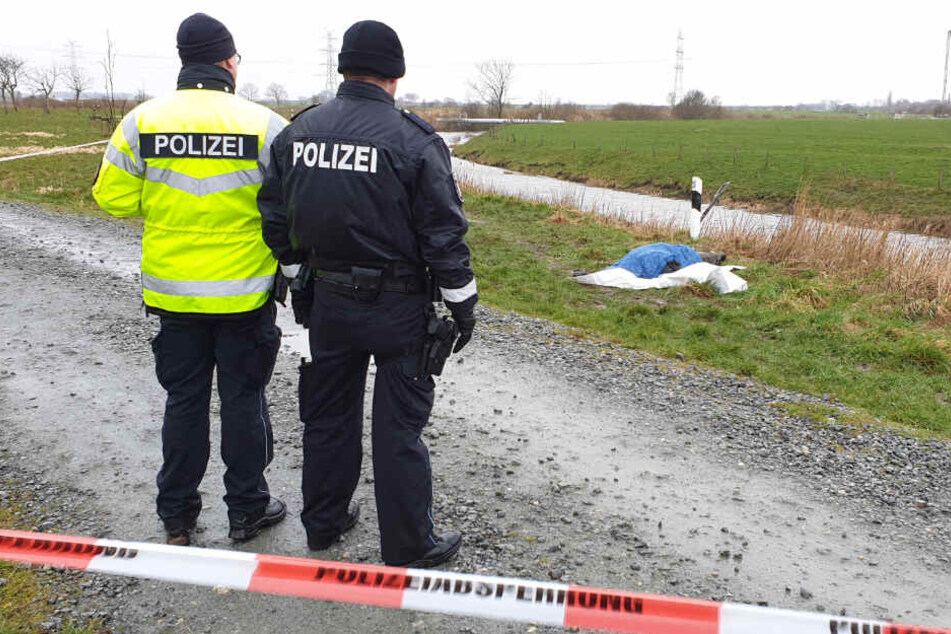 Zwei Einsatzkräfte der Polizei stehen am Fundort: Die Leiche liegt von Tüchern bedeckt im Gras.