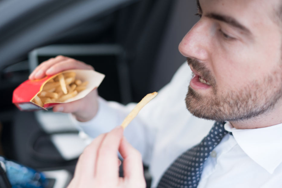 Ein Mann sitzt im Auto und isst ein paar Pommes nebenbei. Die Tüte ist gut gefüllt.