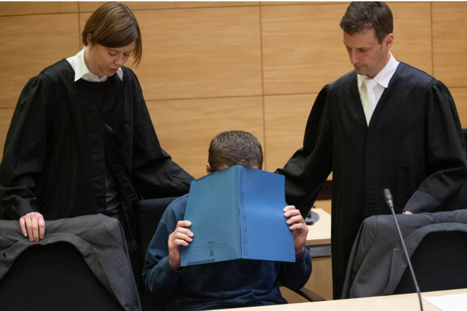 Der Angeklagte sitzt mit einer Mappe vor dem Gesicht im Gerichtssaal neben seinen Verteidigern, Christina Peterhanwahr und Henning Jansen.