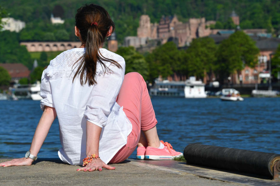 Eine junge Frau sitzt in Heidelberg am Ufer des Neckar und blickt auf die Schlossruine und Teile der Altstadt.