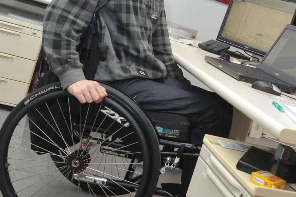 Ein Mann sitzt in einem Rollstuhl an seinem Arbeitsplatz am Schreibtisch.