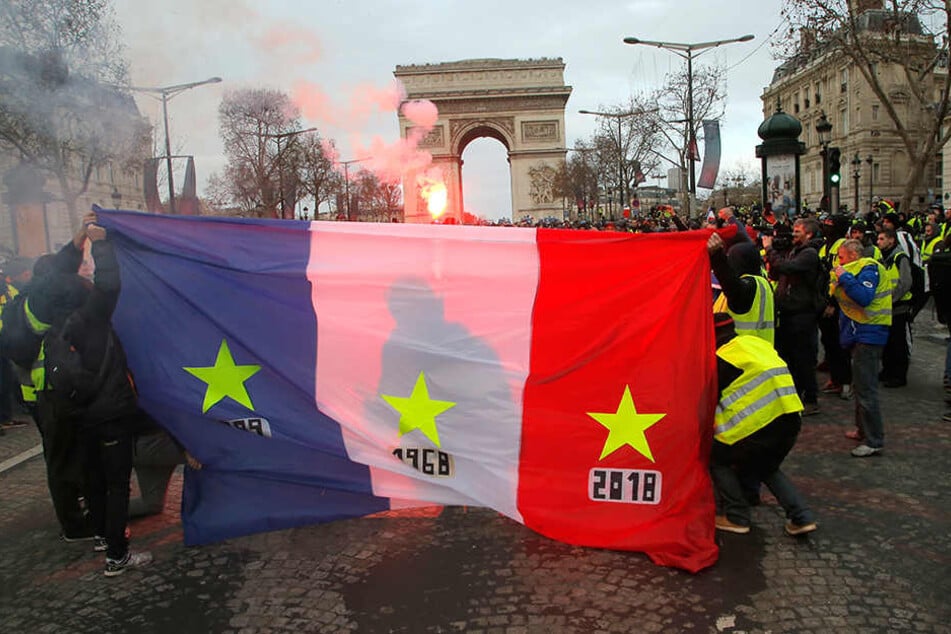 Demonstranten der "Gilets Jaunes", also der "Gelbwesten", halten eine französische Fahne, im Hintergrund ist der Triumphbogen zu sehen.