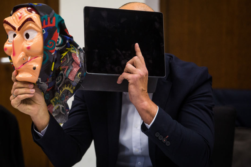 Ein Angeklagter sitzt auf seinem Platz im Amtsgericht, hält sich ein Tablet vor das Gesicht und in der anderen Hand hält er eine Hexenmaske.