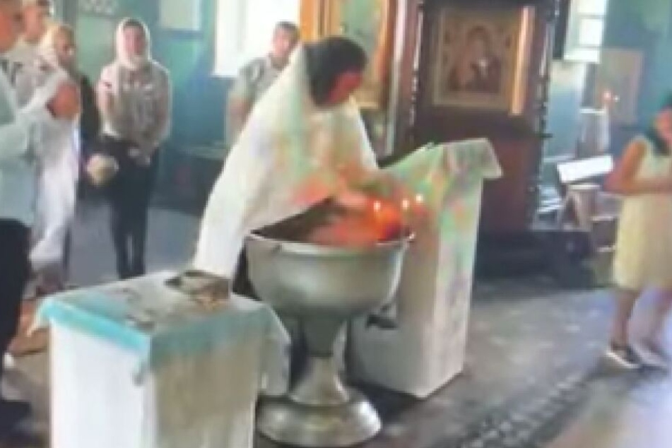Der Priester drückt das schreiende Baby mit der Hand auf seinem Gesicht unter Wasser.