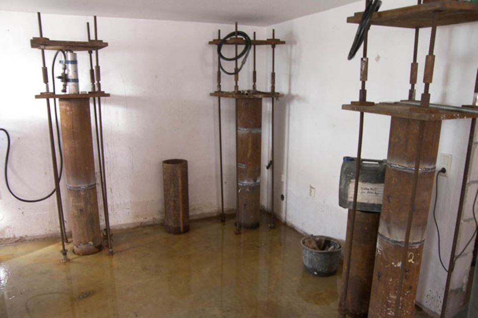 Im Keller werden Pfähle durch die Bodenplatte gebohrt und mit Zugstangen befestigt. Diese werden an der Unterseite verschraubt, können so die Bodenplatte hydraulisch nach oben ziehen.