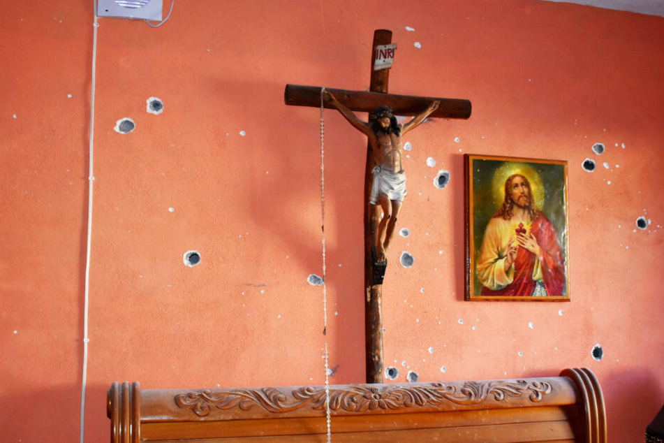 Eine Innenwand eines Hauses, an der ein Kruzifix und ein Bild mit einer Darstellung von Jesus Christus hängen, ist mit Einschusslöchern übersät.