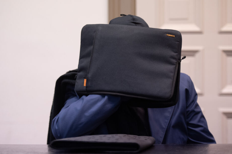 Der Angeklagte versteckt sich hinter einer Tasche.