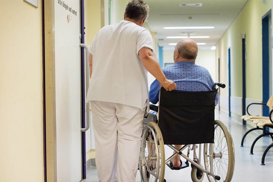 Eine Krankenschwester steht neben einem Patienten im Rollstuhl vor einem Behandlungszimmer.