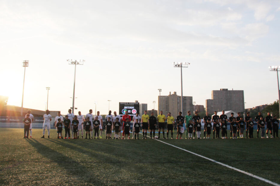 Die Spieler beider Mannschaften - links Cosmos, rechts St. Pauli - stehen im Abendlicht mit Kindern und den Schiedsrichtern auf dem Platz.