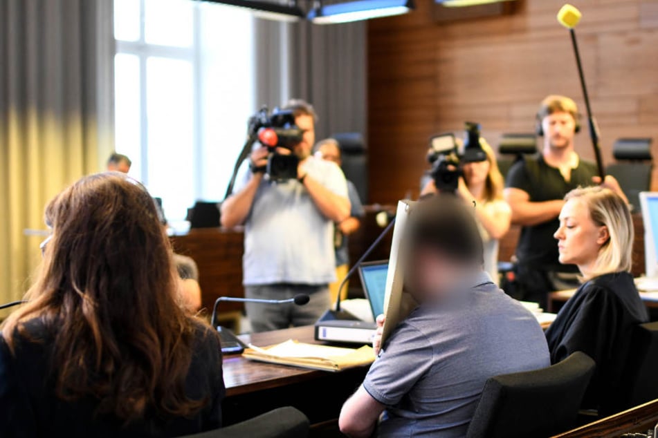 Der Angeklagte (2.v.r.) hält sich im Gerichtssaal einen Aktendeckel vors Gesicht.