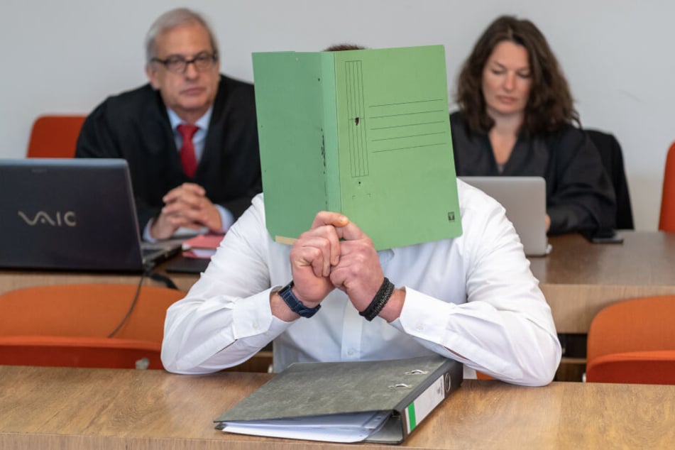 Der Angeklagte hält sich vor Prozessbeginn einen Aktendeckel vor das Gesicht.