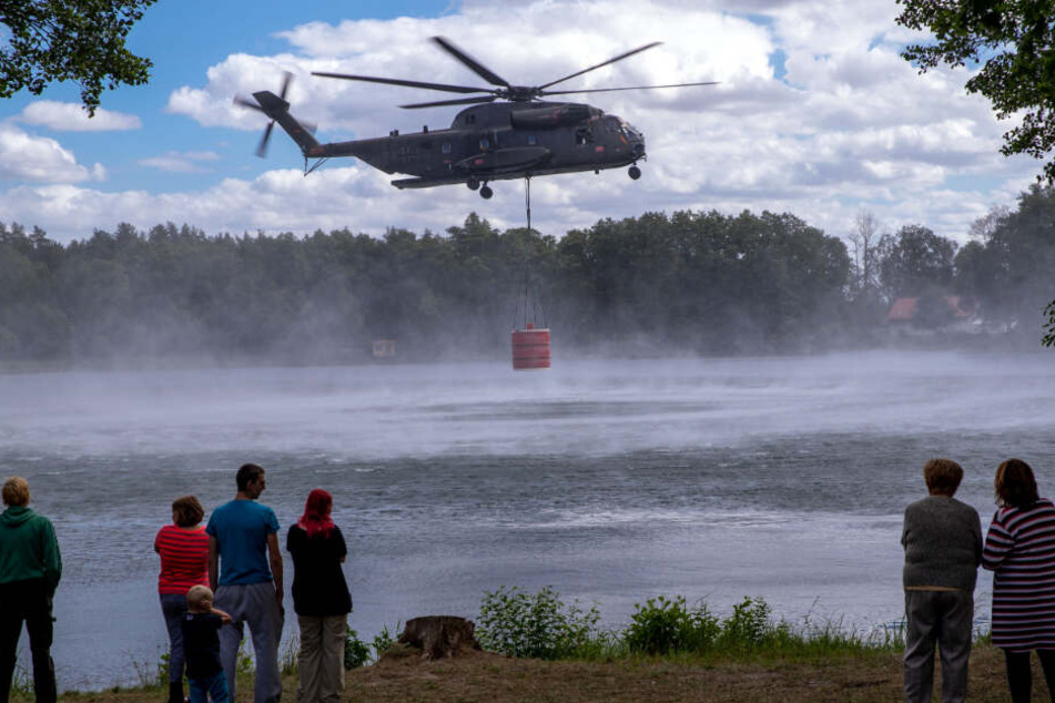 Hubschrauber fliegen mit einer großen Wasserladung Richtung Waldbrand. Schaulustige gucken gespannt zu.