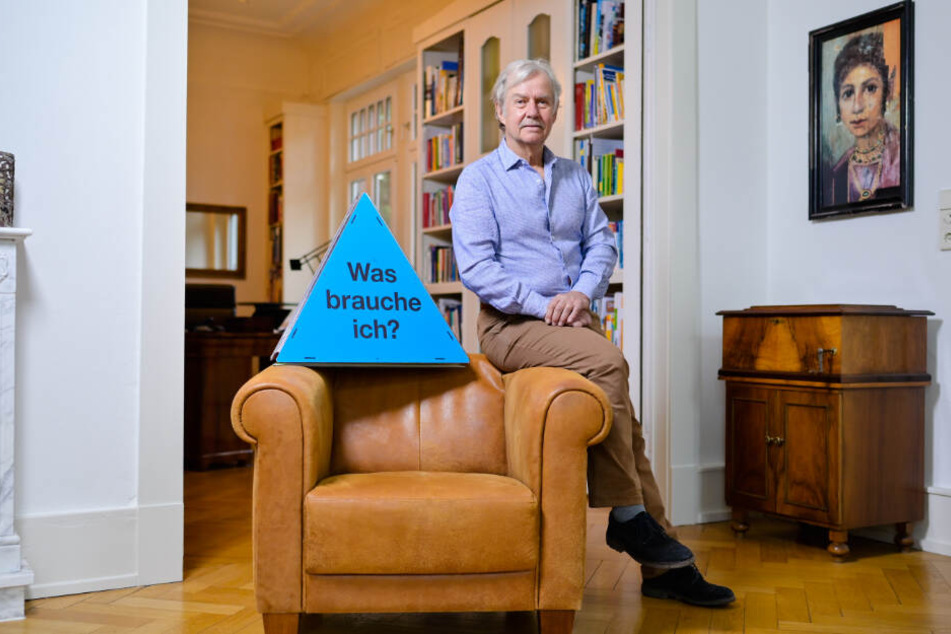 Ernst Fritz-Schubert sitzt auf einer Sessellehne, neben ihm steht ein Dreieck mit der Aufschrift "Was brauche ich"
