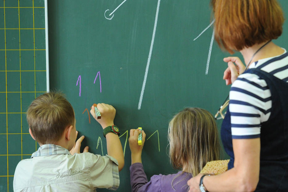 Unter Anleitung der Klassenlehrerin schreiben zwei Grundschüler die Zahl "Eins" an die Tafel.