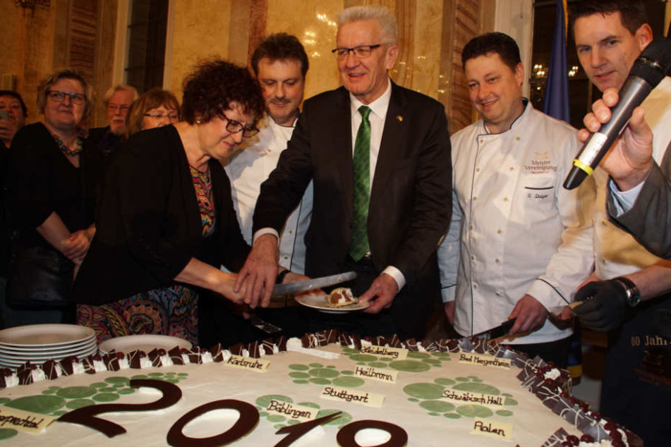 Winfired Kretschmann (Mitte) schneidet gemeinsam mit seiner Frau Gerlinde (links) eine Torte in Form des Bundeslandes Baden-Württemberg an.