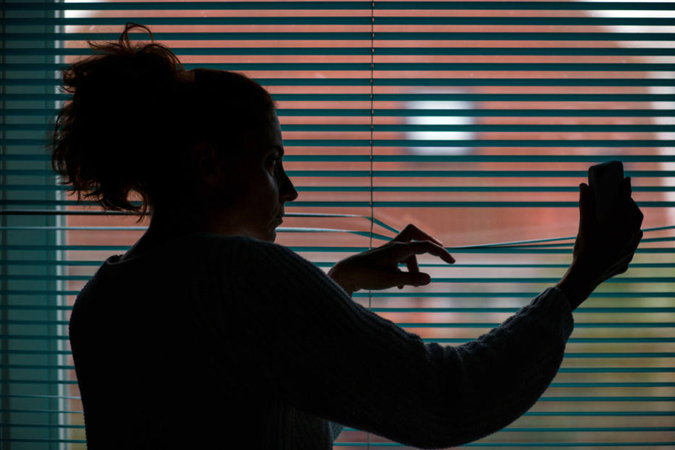 Eine Frau beobachtet durch eine geschlossene Jalousie nach einem Stalking-Anruf die Straße vor ihrem Haus. (Symbolbild)