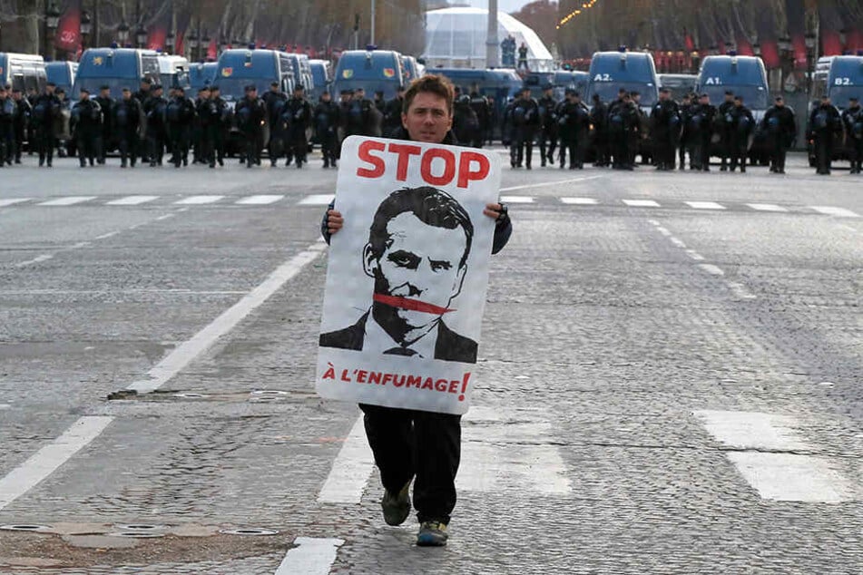 Ein Demonstrant mit einem Plakat mit dem Abbild des französischen Präsidenten geht auf der Champs Elysees, im Hintergrund stehen Polizisten.