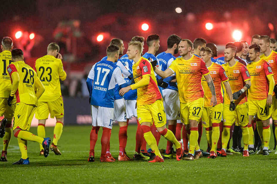 Die Mannschaften begrüßen sich vor dem Anpfiff. Im Hintergrund zünden Fans Pyrotechnik.