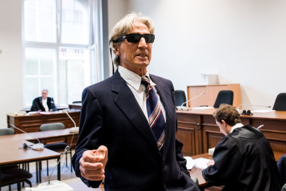 Der Angeklagte steht mit Sonnenbrille auf der Nase und im Anzug im Gerichtssaal.