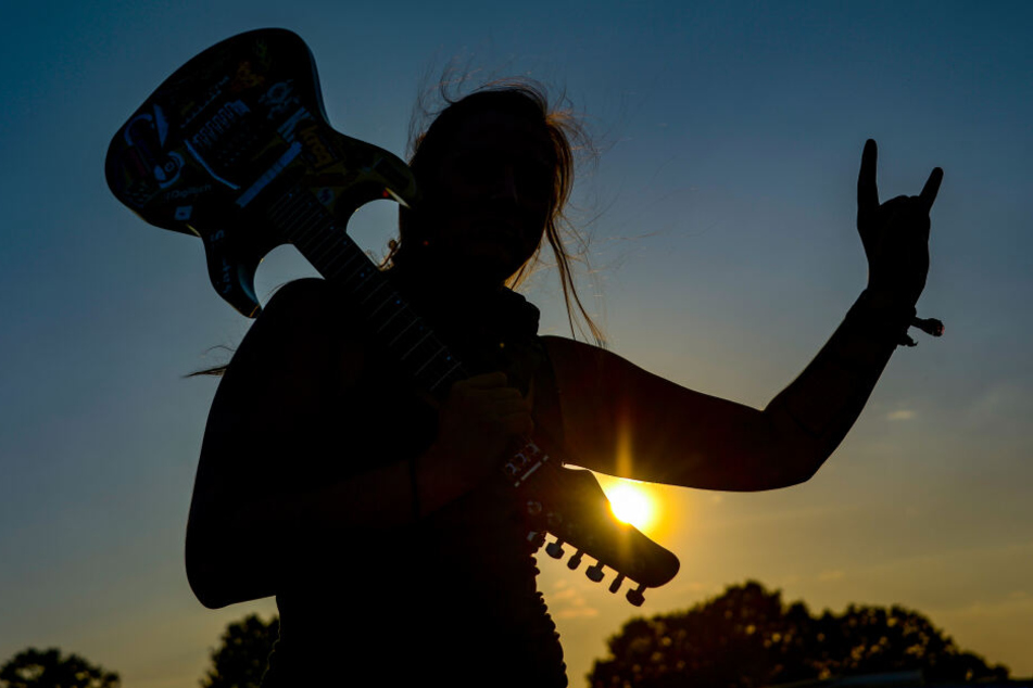 Eine Frau mit einer E-Gitarre auf der Schulter zeigt beim Wacken Open Air auf dem Festivalgelände den Metalgruß, die sogenannte "Pommesgabel".