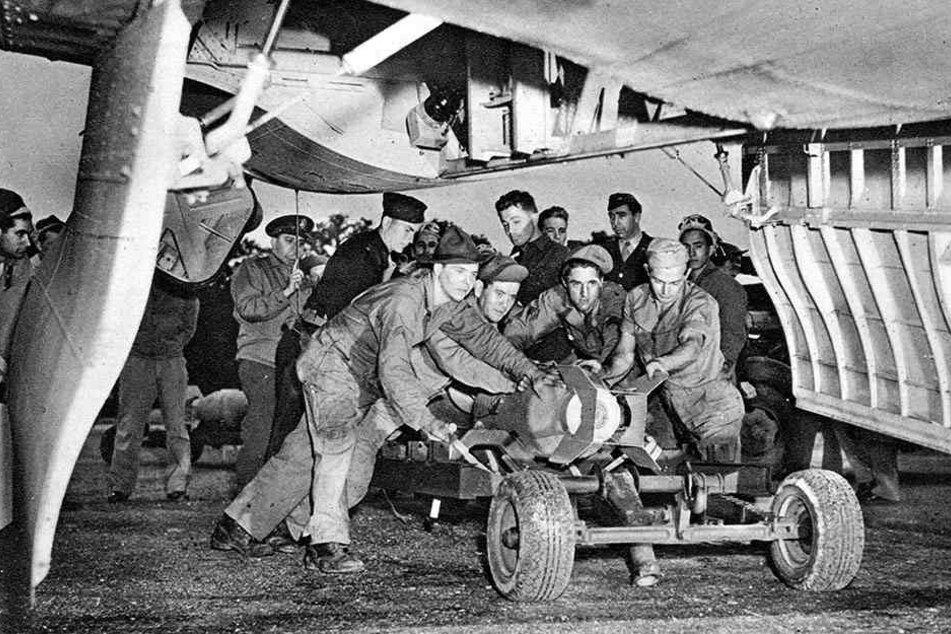 Bodentruppen rollen einen Wagen mit einer Bombe unter ein Kampfflugzeug in Position.