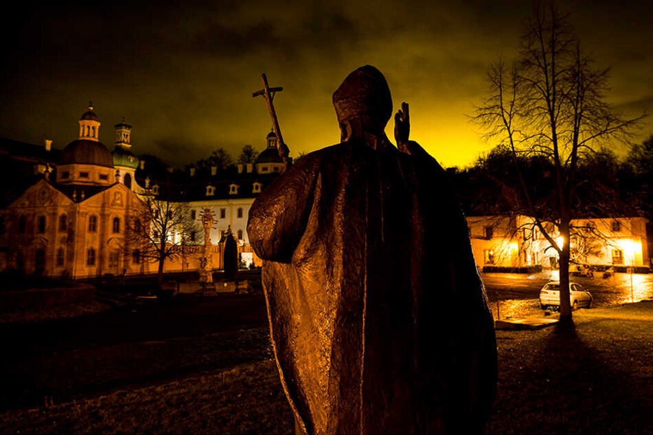 Eine Bronzestatue vor dem Kloster St. Marienthal erinnert an Johannes Paul II. Durch die Beleuchtung der Tomatenreiferie in Bogatynia, Polen, ist zu den Abendstunden ein heller Schein zu sehen.