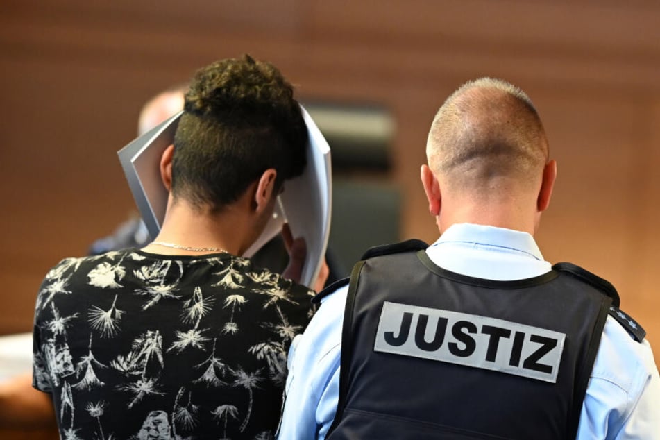 Einer von elf Angeklagten wird vor dem Prozessbeginn von einem Justizbeamten in einen Gerichtssaal im Landgericht gebracht. (Archivbild)