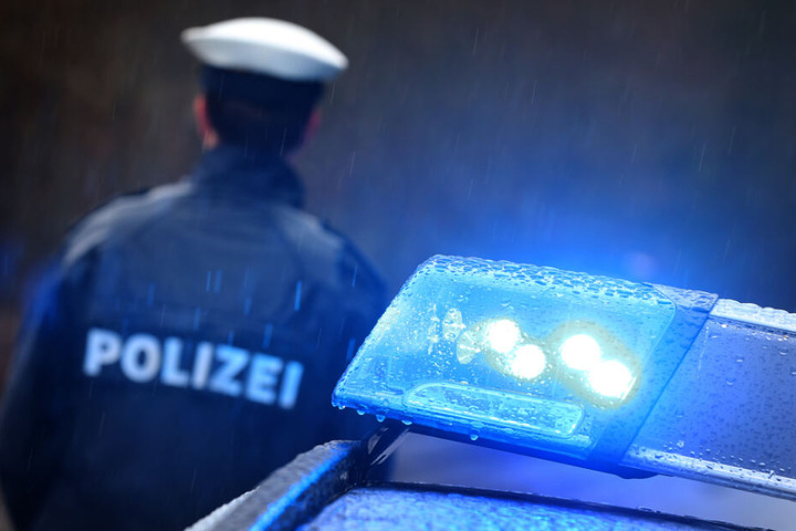 Ein Polizist steht im Regen vor einem Streifenwagen dessen Blaulicht aktiviert ist (Symbolbild).