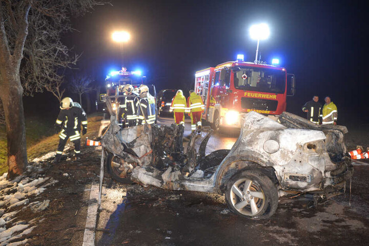 Feuerwehrleute und Rettungskräfte stehen auf einer Landstraße hinter dem ausgebrannten Auto.