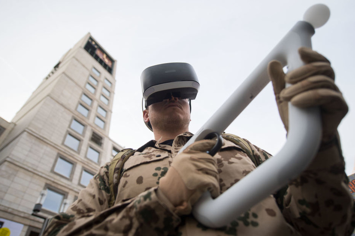 Ein Rüstungsgegner in Uniform hat vor dem Rathaus bei einer Kundgebung von Rüstungsgegnern gegen die Militärmesse ITEC eine Konsolenspielzeugwaffe in den Händen und eine VR-Brille auf dem Kopf.