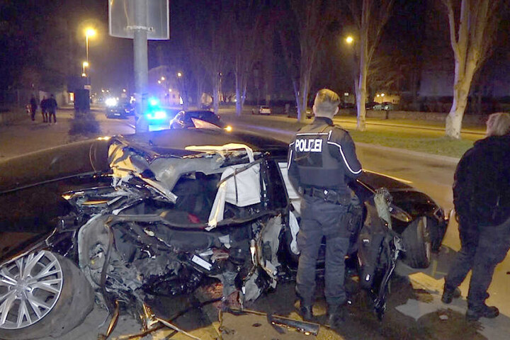 Autorennen auch in Magdeburg? Polizisten stehen an einem völlig demolierten Auto. Nach dem Verkehrsunfall ermittelt die Polizei, ob es möglicherweise vorher ein illegales Autorennen gegeben hat.