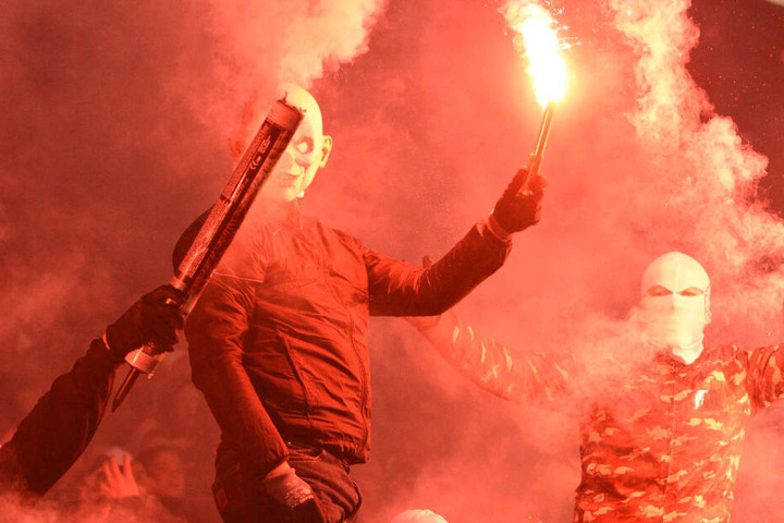 Mit Masken und vermummt halten Fans Pyro-Technik hoch.