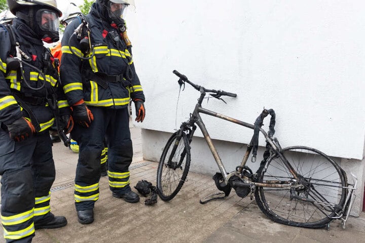 Zwei mit Atemschutzmasken bekleidete Feuerwehrmänner gucken sich das zerstörte E-Bike an.