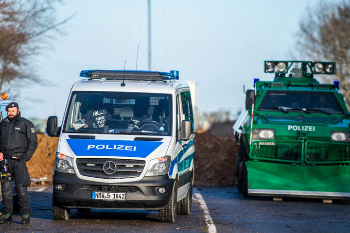 Polizisten stehen im Hambacher Forst auf einer Straße neben Polizeiautos und einem Räumpanzer.