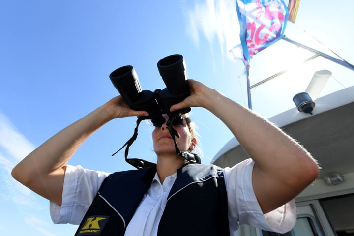 Eine Beamtin der Wasserschutzpolizei steht mit einem Fernglas am Heck eines Bootes. (Archivbild)