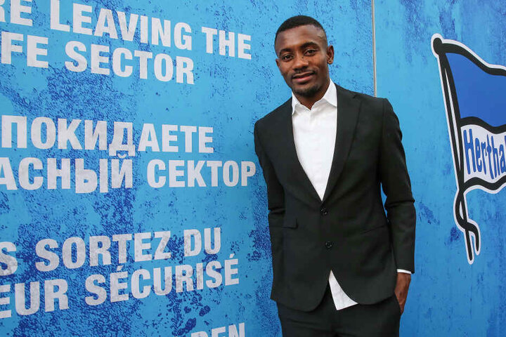 Salomon Kalou steht lächelnd an Wand in der Messehalle mit Vereinslogo und Schriftzug: