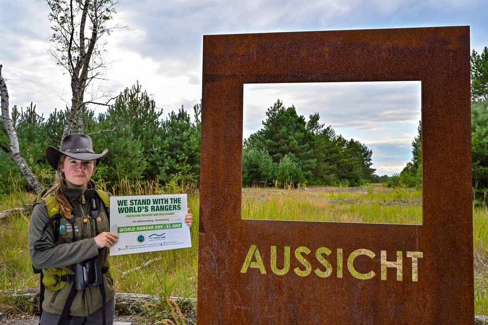Sophie Büchner ist Rangerin, steht in der Lieberoser Heide und zeigt ein Plakat zum World Ranger Day. Die junge Frau arbeitet bei der Stiftung Naturlandschaften Brandenburg.