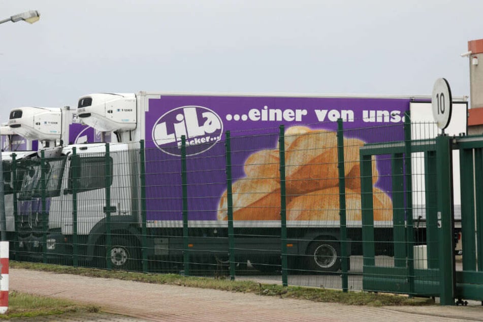 Lastwagen von "Lila Bäcker" stehen am Produktionsstandort hinter einem Zaun.