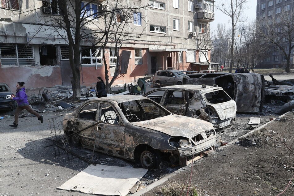 Menschen gehen an zerstörten Autos und Trümmern in Mariupol vorbei. Frankreich will mit der Türkei und Griechenland eine humanitäre Aktion in Gang setzen, um kurzfristig Menschen aus der schwer umkämpften ostukrainischen Hafenstadt zu retten.