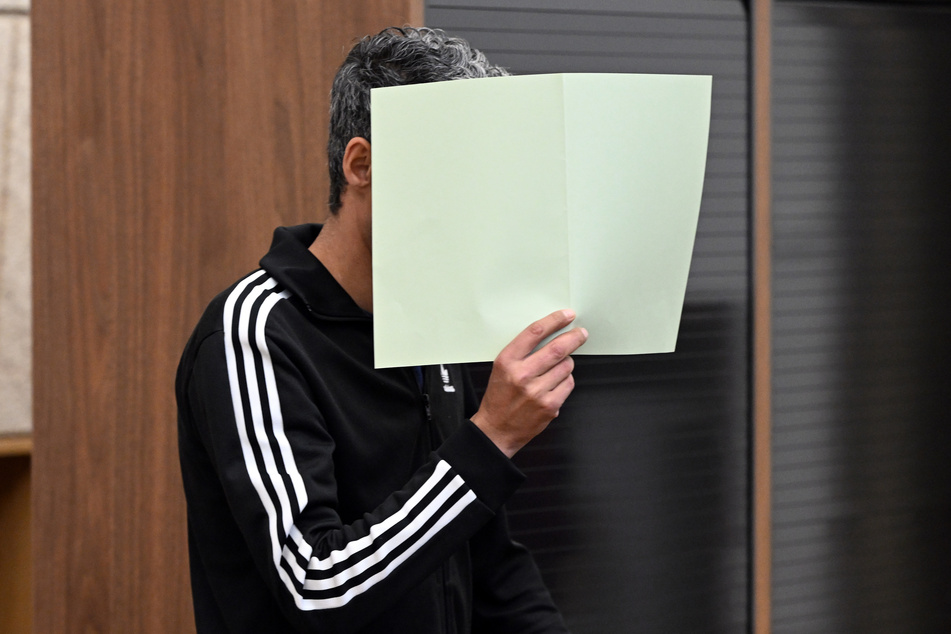 Der Angeklagte hält sich im Gerichtssaal eine grüne Mappe vor das Gesicht.
