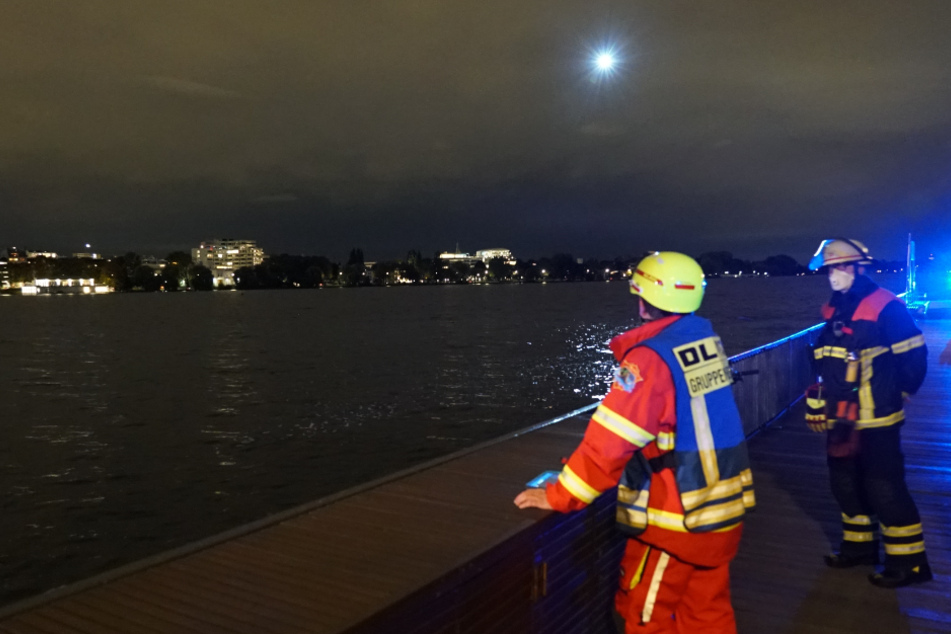 Einsatzkräfte stehen am Ufer, während ein Hubschrauber das Wasser absucht.