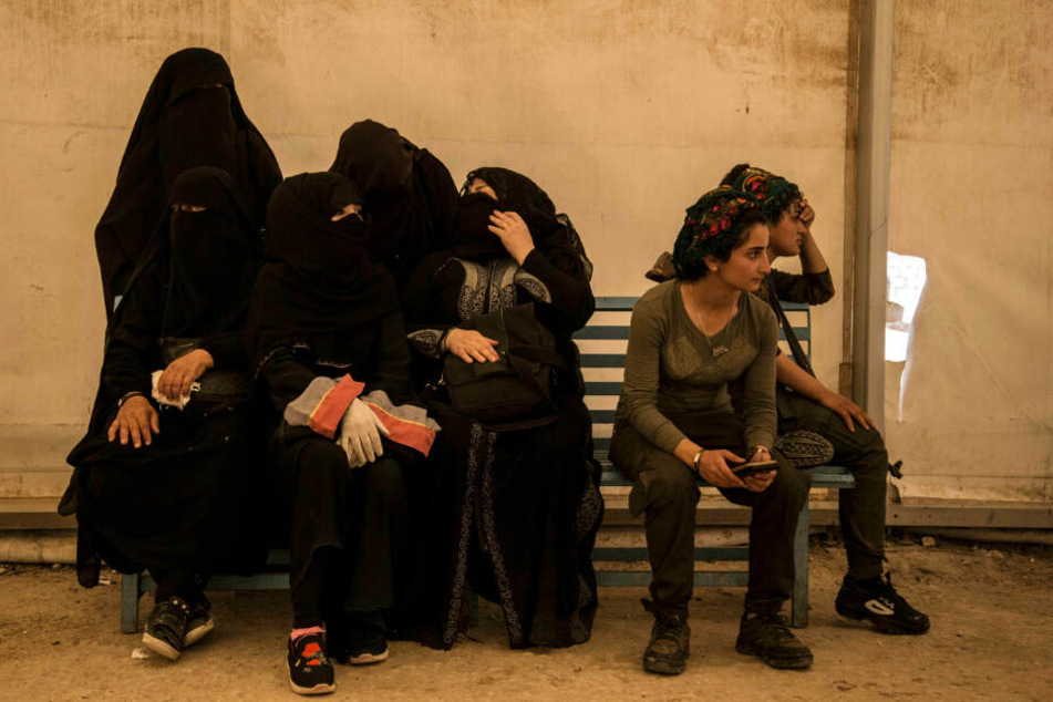 Frauen (l), die mit Kämpfern der Terrormiliz Islamischer Staat (IS) verwandt sind, sitzen im Flüchtlingslager Al-Hol in der Provinz Hasakeh, Syrien, neben Wächter der Syrischen Demokratischen Kräfte (SDF), während sie darauf warten, in Busse und Lastwage